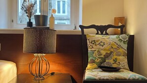 Une pièce avec une table d’appoint en bois, une lampe, une chaise avec un coussin à motifs floraux et une fenêtre donnant sur des immeubles.