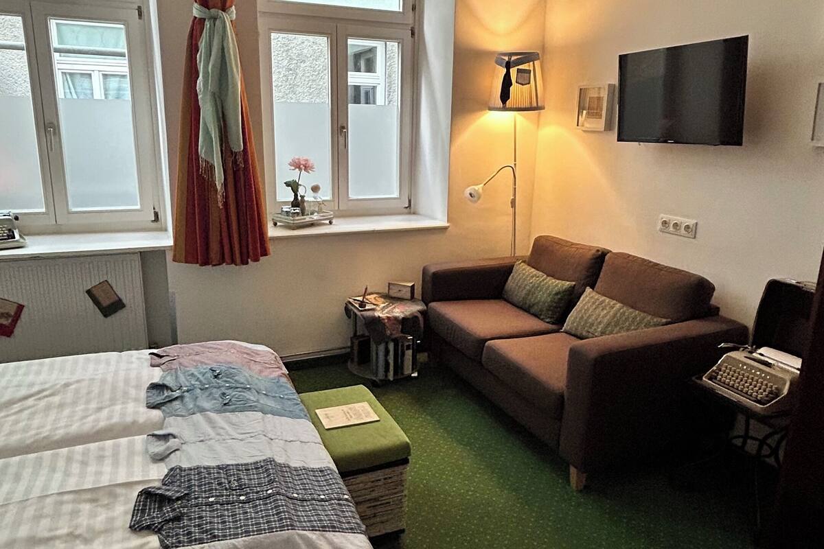 Una habitación con cama, sofá, escritorio con máquina de escribir, lámpara y televisión en la pared.