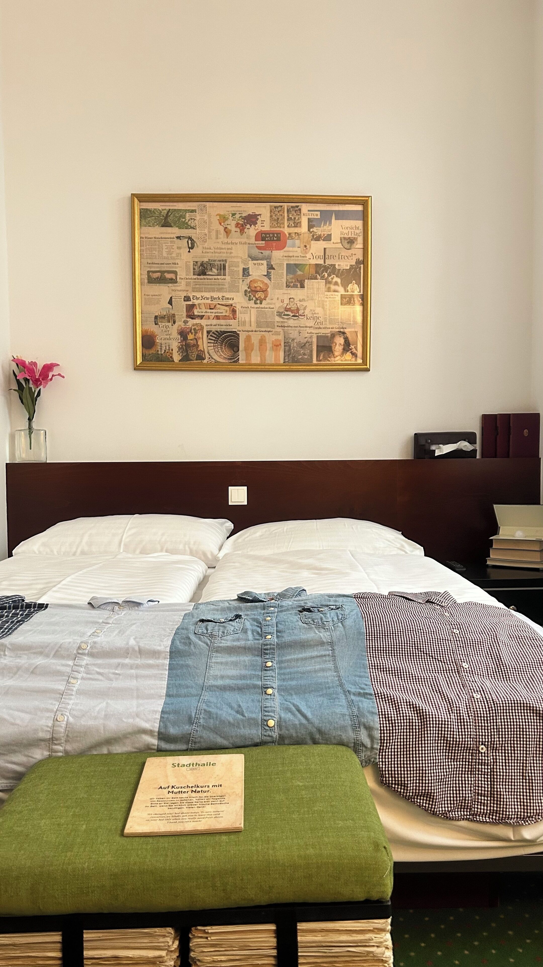 Habitación de hotel con un gran collage enmarcado en la pared, una cama con almohada verde y una mesita de noche con un libro.