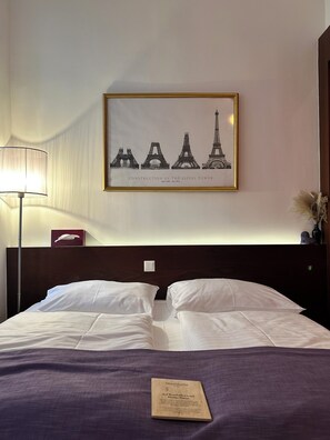A hotel room with a bed, a framed picture of the Eiffel Tower, a lamp, and a small decorative item on the nightstand. - Pension Stadthalle (Vienna)