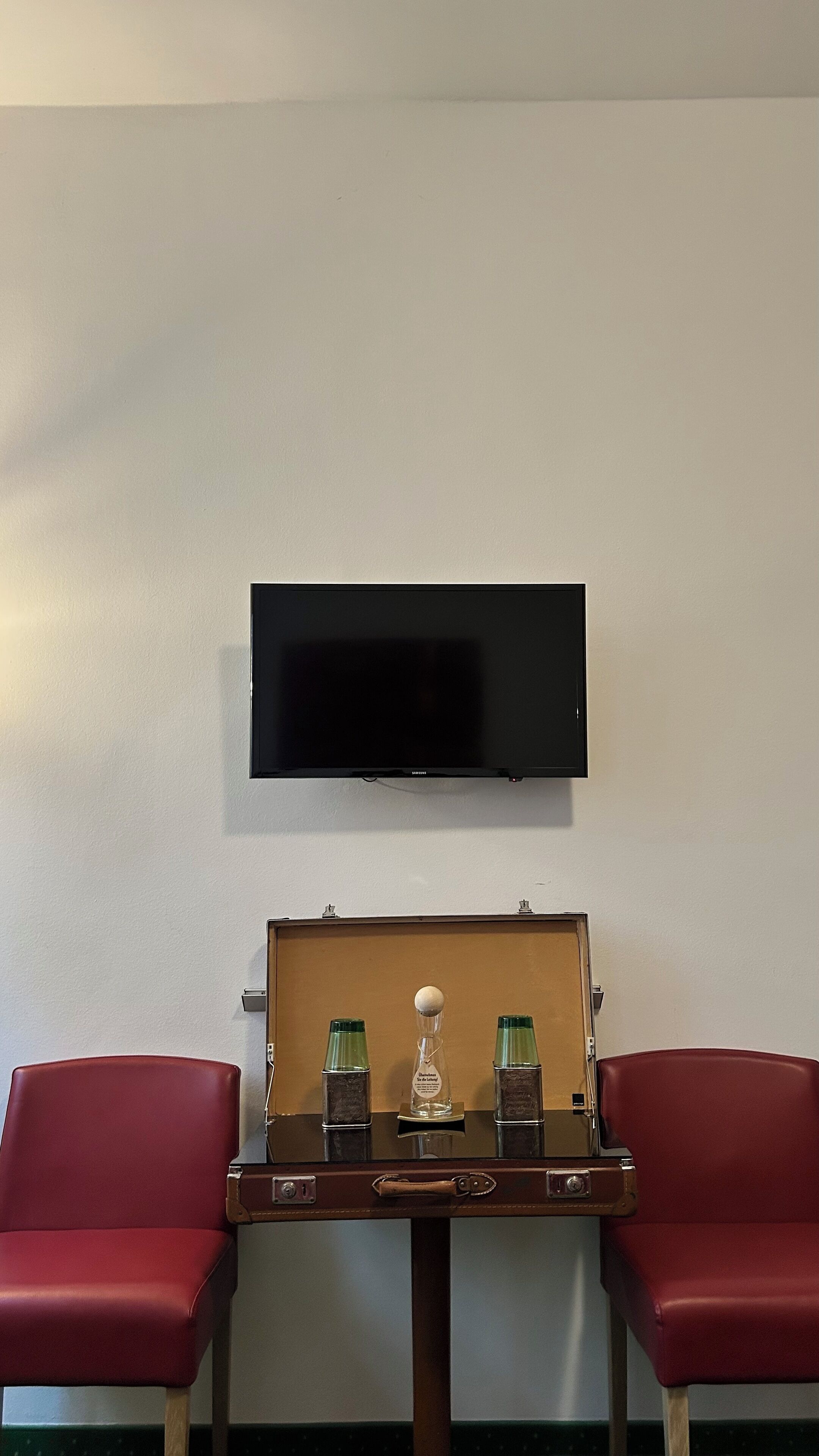 A room with a flat-screen TV mounted on the wall, a vintage suitcase table with two red chairs, and a small decorative item on the table.