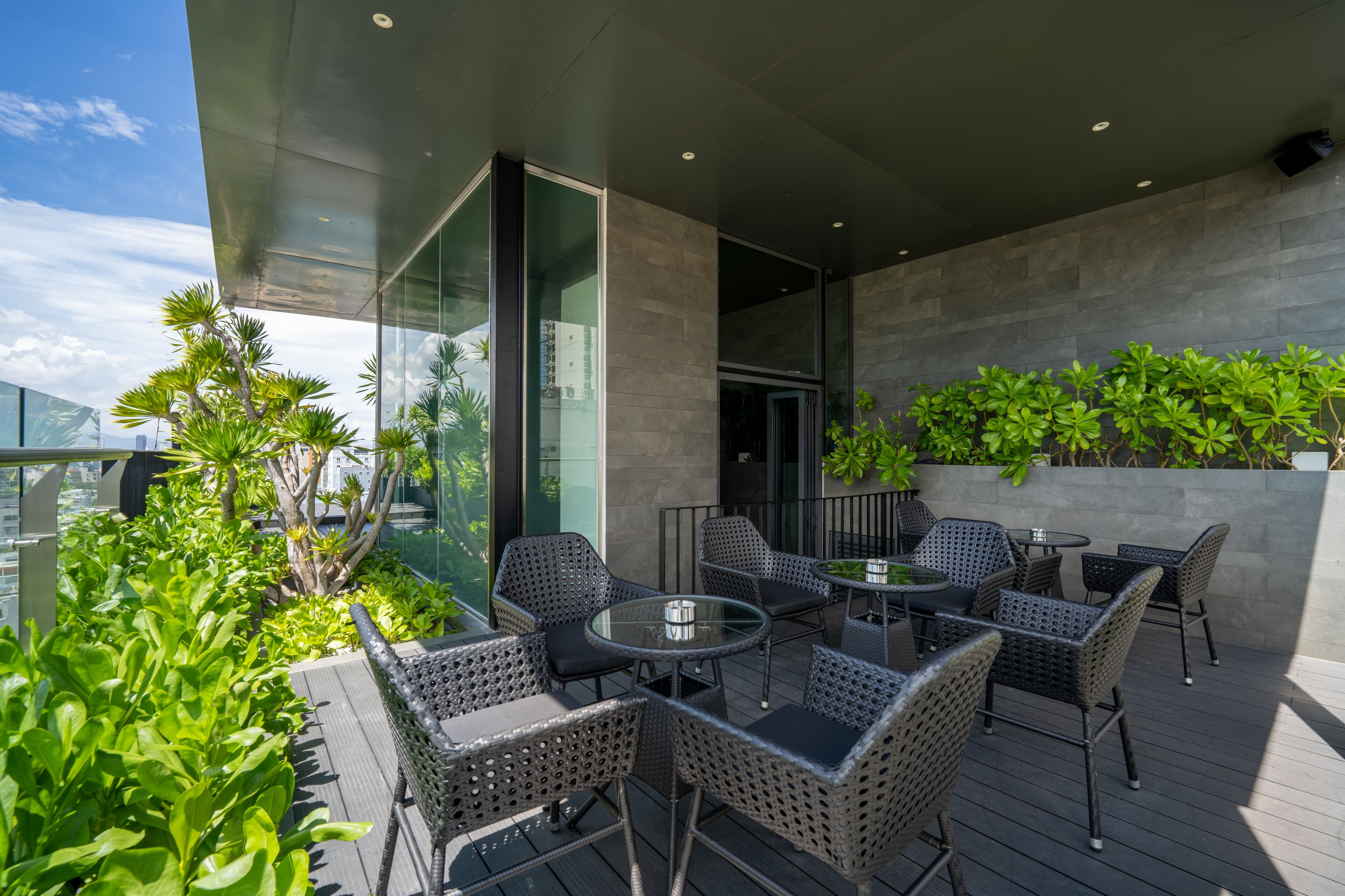 A modern outdoor patio with wicker furniture and glass tables, surrounded by lush greenery and a cityscape view.