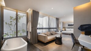 A modern hotel room with a large bathtub, a sofa, a dining table, and a view of the city. - LEGEND Boutique Hotel (Da Nang)