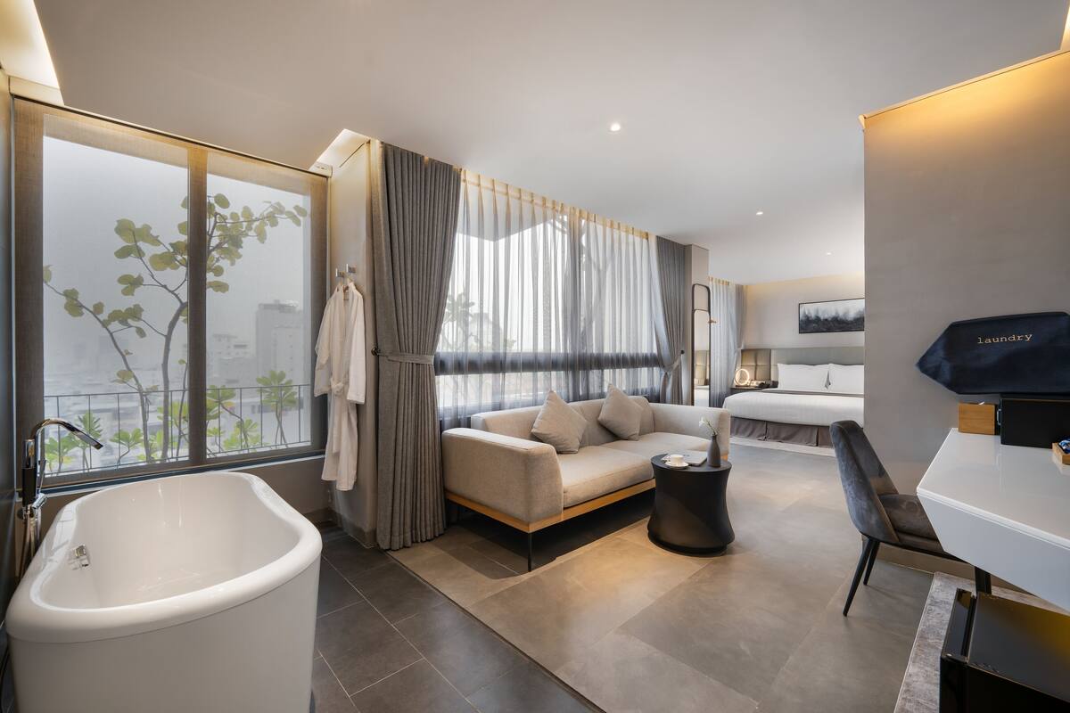 A modern hotel room with a large bathtub, a sofa, a dining table, and a view of the city.