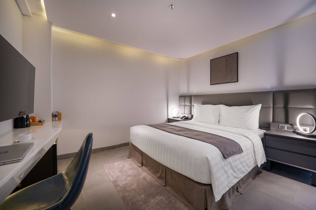 A modern hotel room with a neatly made bed, a desk with a laptop, a chair, and a bedside table with a mirror.