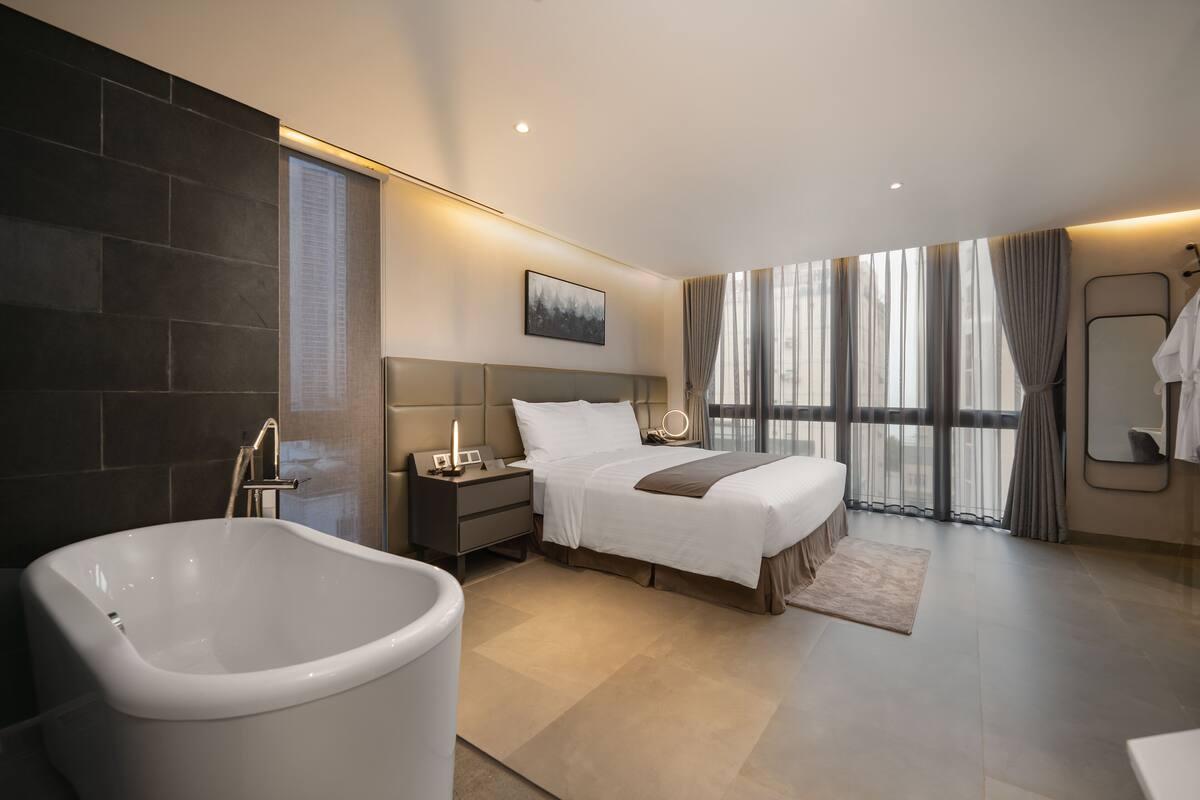 A modern hotel bedroom with a large bathtub, a neatly made bed, a bedside table with a lamp, and a large window with curtains.