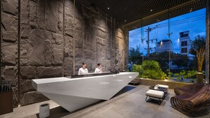 A reception desk with a marble top, two staff members, a wicker chair, and a view of a building through a glass wall. - LEGEND Boutique Hotel (Da Nang)