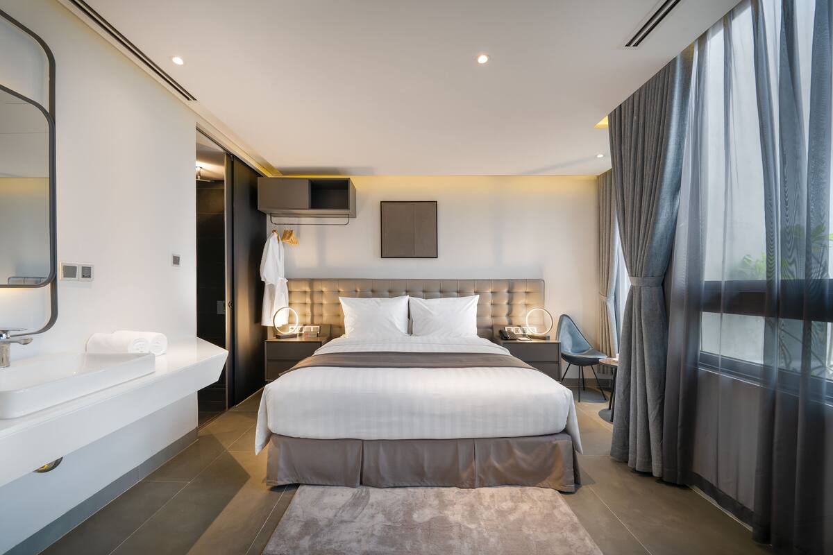 A modern hotel room with a large bed, a separate bathroom with a vanity, a flat-screen TV, and a large window with curtains.