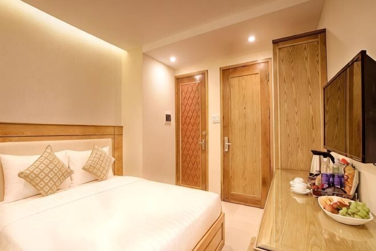 A hotel room with a bed, wooden wardrobe, and a table with toiletries and fruit.