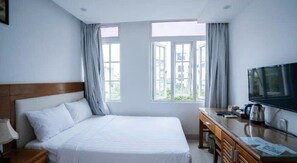 A hotel room with a bed, a desk with a TV, and two windows with curtains.