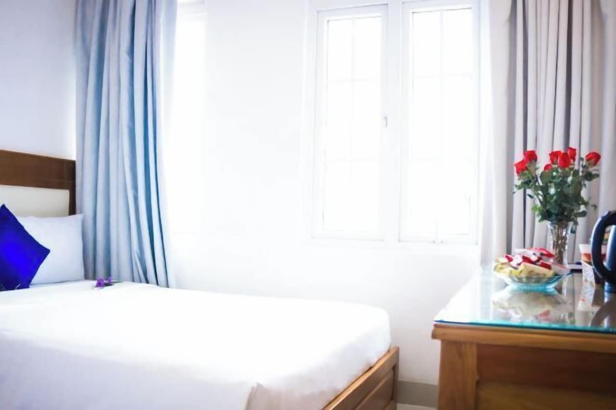 A hotel room with a bed, a desk, a vase with flowers, and a window with curtains.