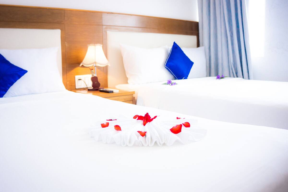 A hotel room with two beds, each with white bedding and blue pillows. There is a nightstand with a lamp and a remote control.
