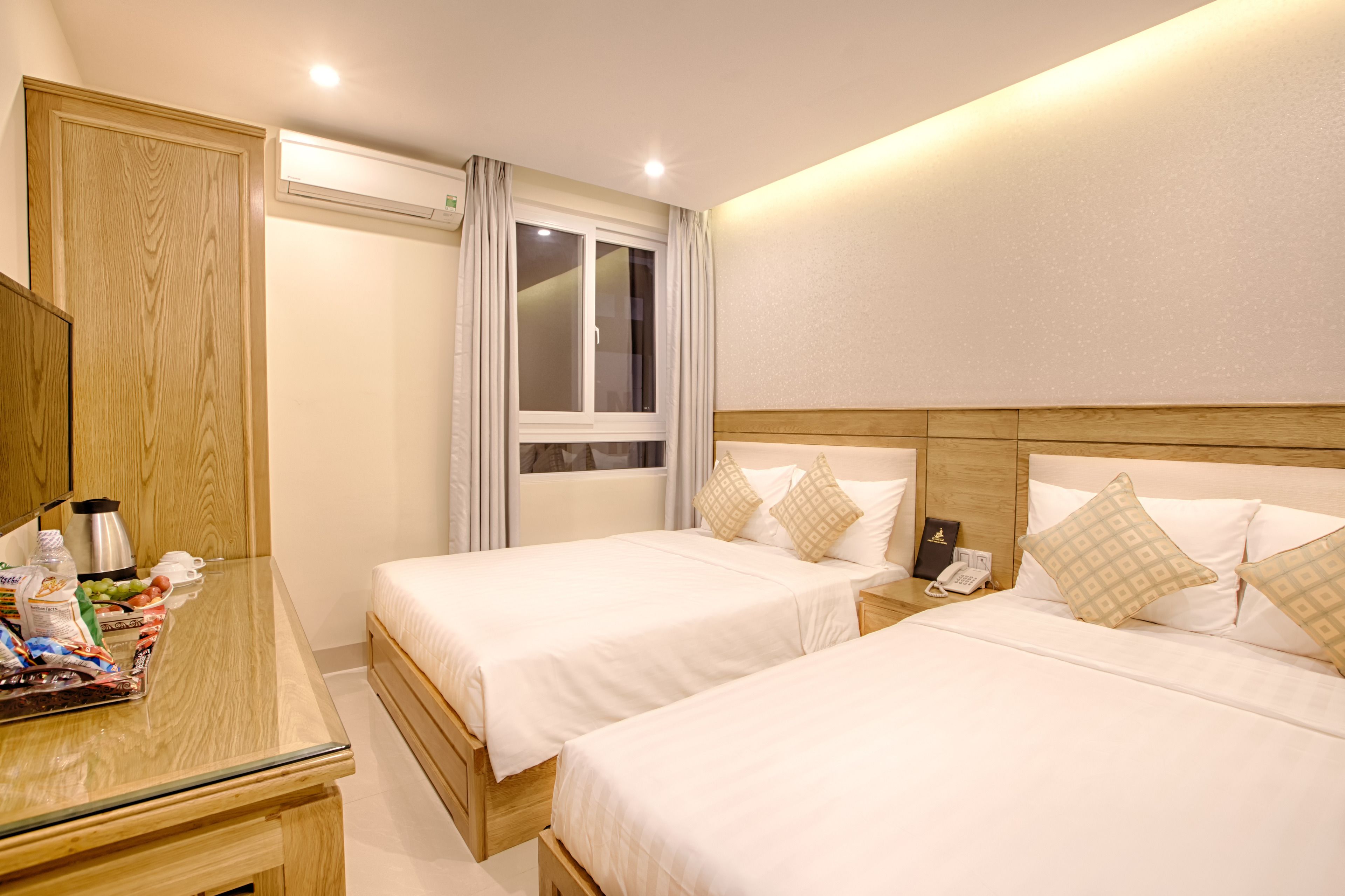 A hotel room with two beds, a wooden wardrobe, a bedside table with a lamp, and a window with curtains.