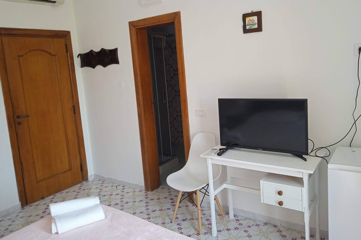 Una habitación con puerta de madera, un escritorio blanco con televisor de pantalla plana, una silla blanca y un pequeño marco de fotos en la pared.