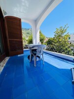 Un balcón con piso de baldosas azules, una mesa con mantel blanco y una silla.