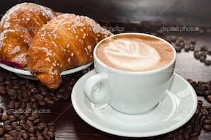 Un croissant y una taza de café con arte latte sobre una superficie de madera, rodeado de granos de café.