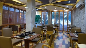 Un restaurant au décor moderne, avec un mélange de meubles en bois et de meubles rembourrés, de grandes fenêtres équipées de stores et un sol à motifs.