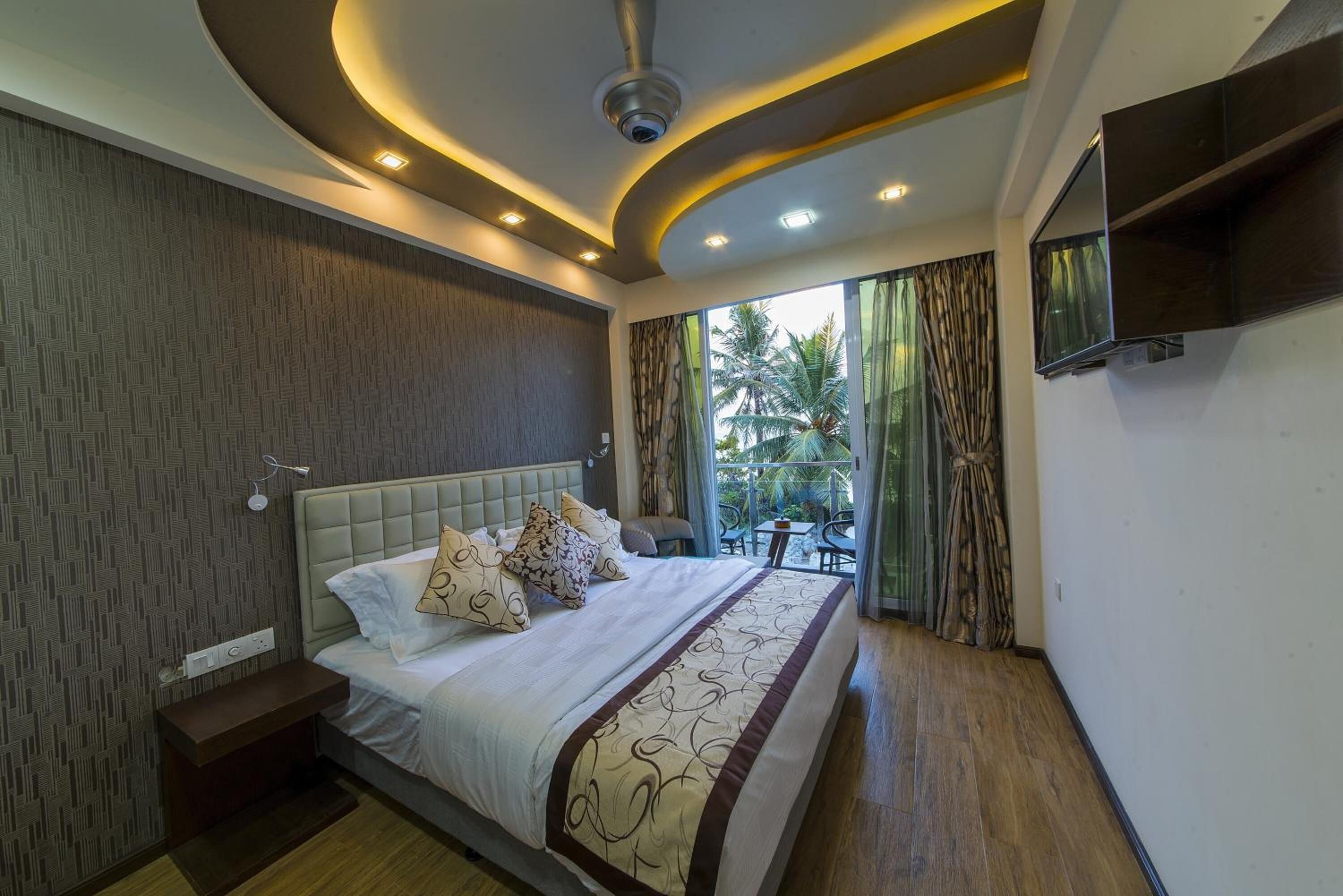 Une chambre moderne avec un grand lit, un ventilateur de plafond, un éclairage encastré, une télévision et une vue sur l’extérieur.
