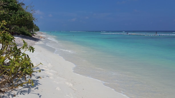 Une plage avec une eau turquoise et limpide, du sable blanc et quelques bateaux visibles au loin.