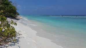 Una playa con agua turquesa cristalina, arena blanca y algunos barcos a lo lejos.