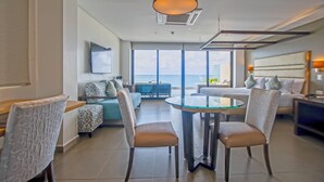 Une chambre d’hôtel moderne avec une table à manger en verre, deux chaises, un canapé, un lit avec une tête de lit et une grande fenêtre donnant sur la mer.