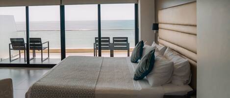 A hotel room with a large bed, a view of the sea, and a balcony with chairs.