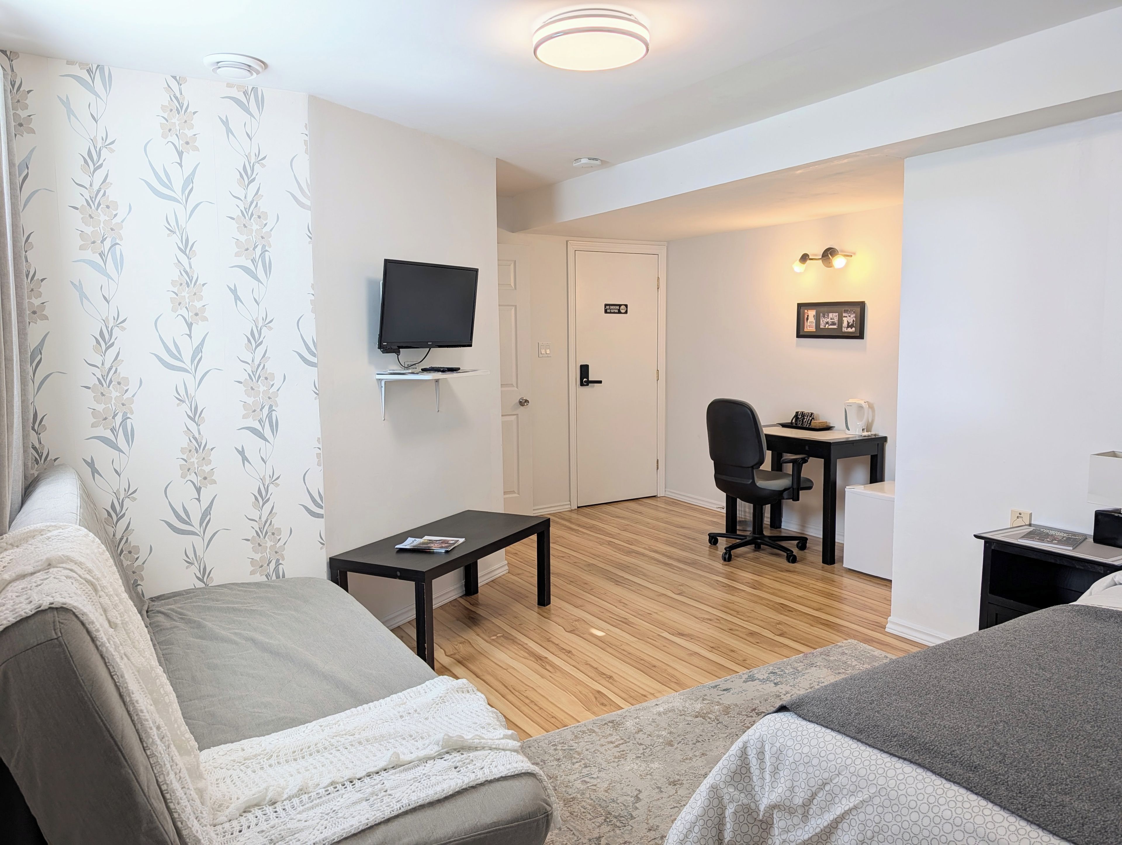 A room with a television, a desk with a chair, a small table, and a bed with a blanket.