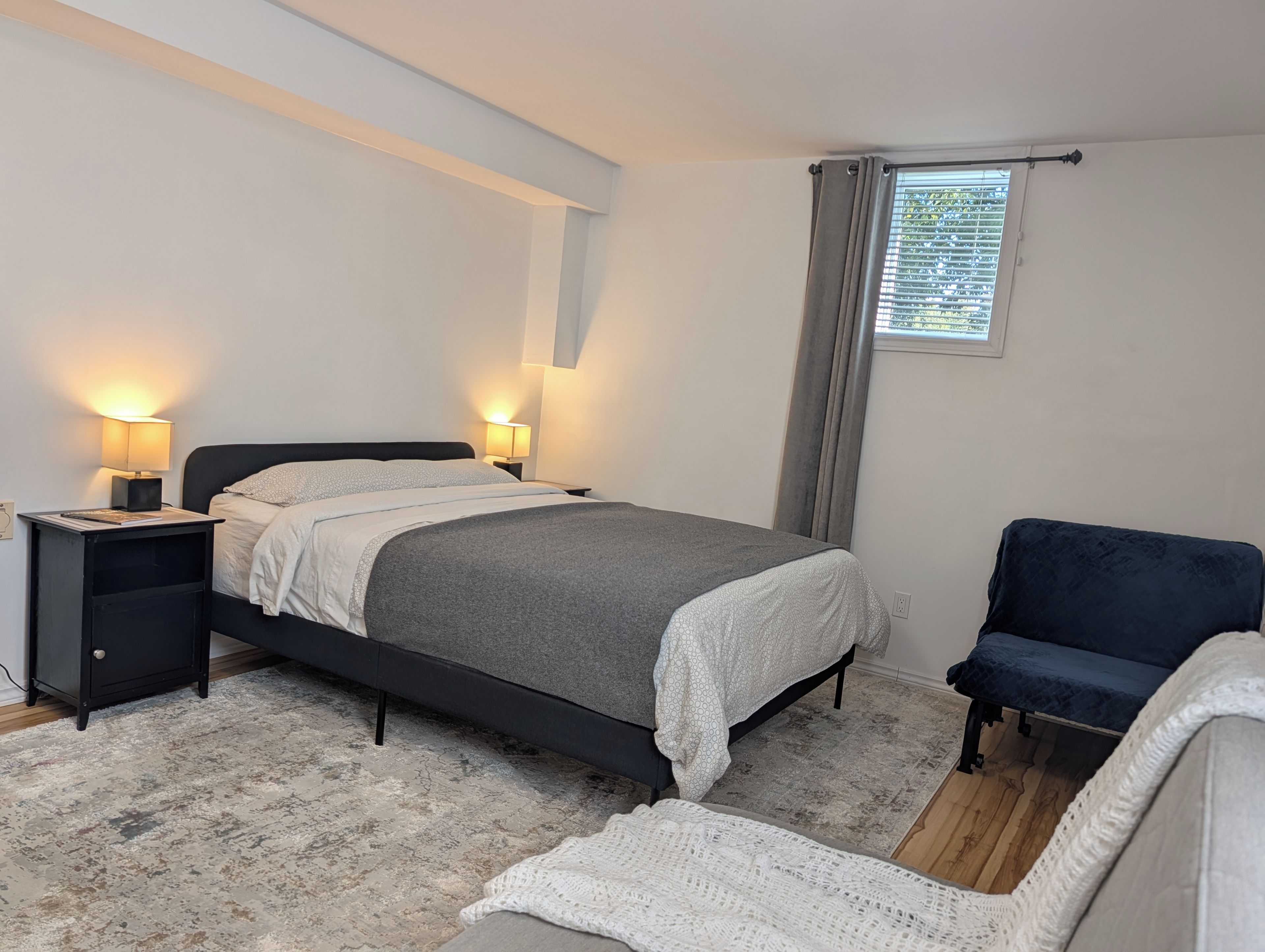 Un dormitorio con cama, mesitas de noche, una silla y ventana con cortinas.