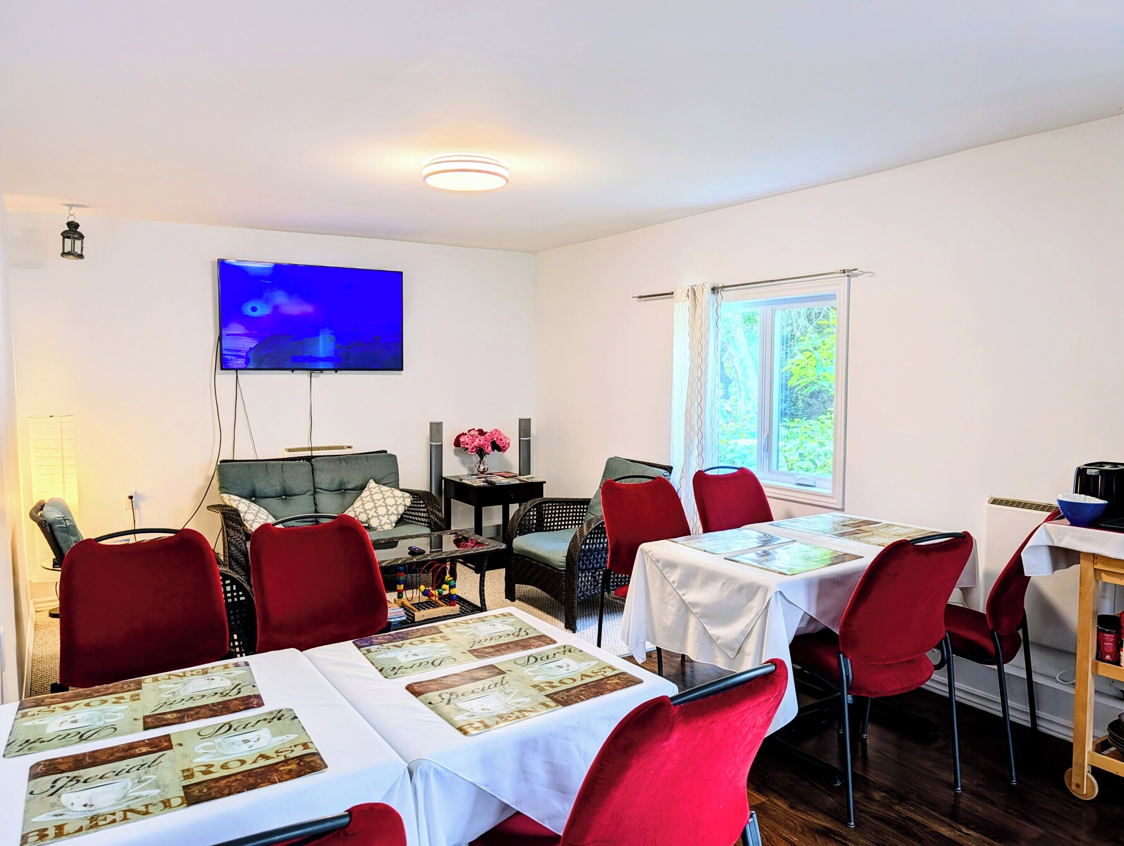 Un comedor con una mesa preparada para dos personas, sillas rojas, un televisor montado en la pared y una ventana con cortinas.