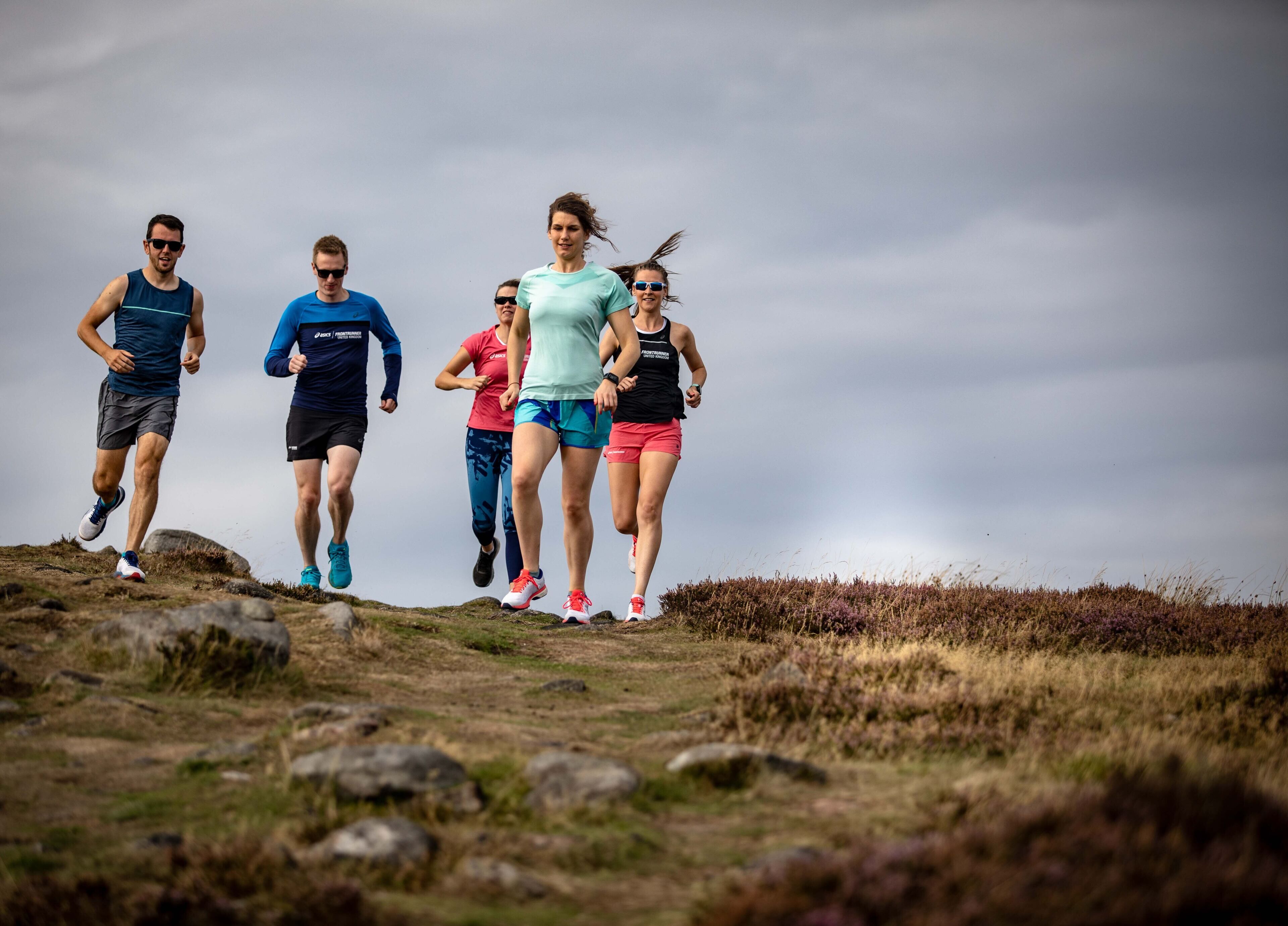 A group of people running on a grassy hill with a cloudy sky in the background.