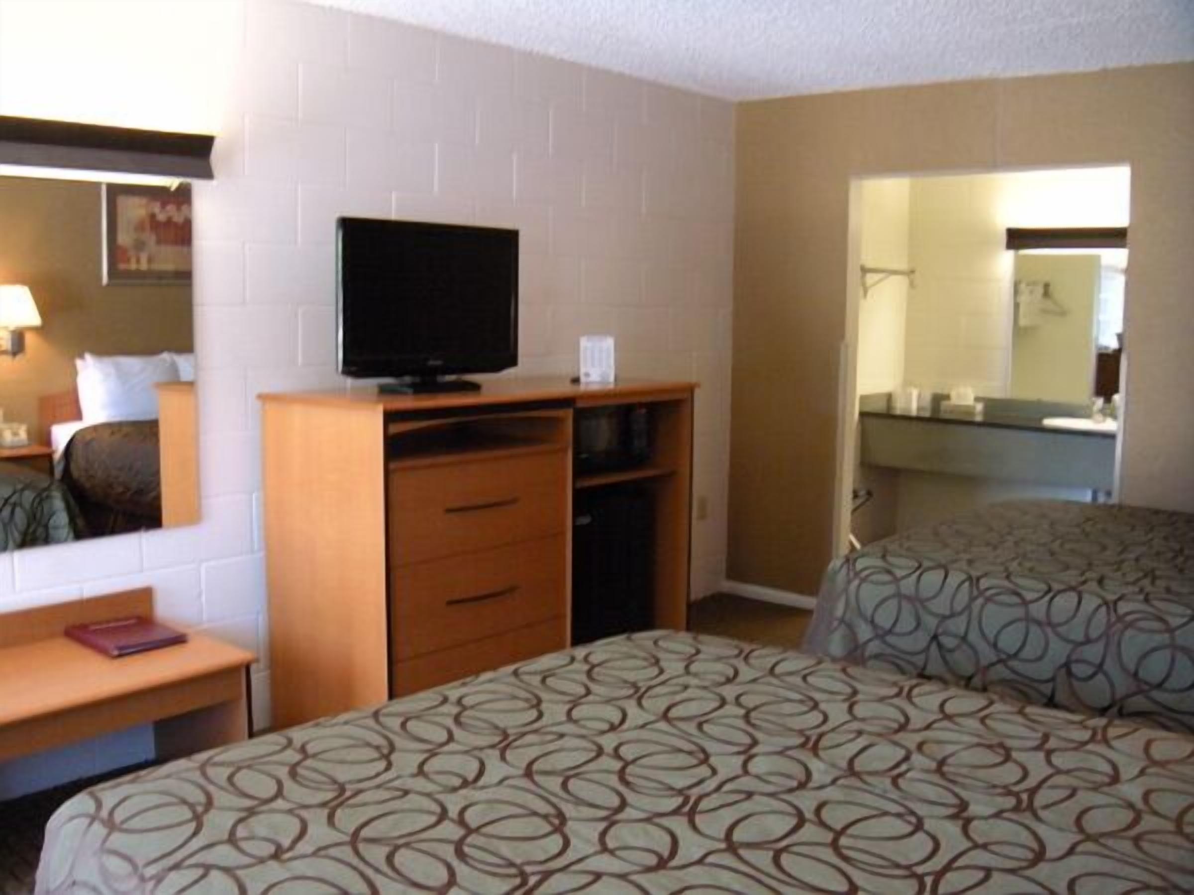 A hotel room with two beds, a TV on a wooden dresser, a desk with a lamp, and a bathroom visible through an open door.