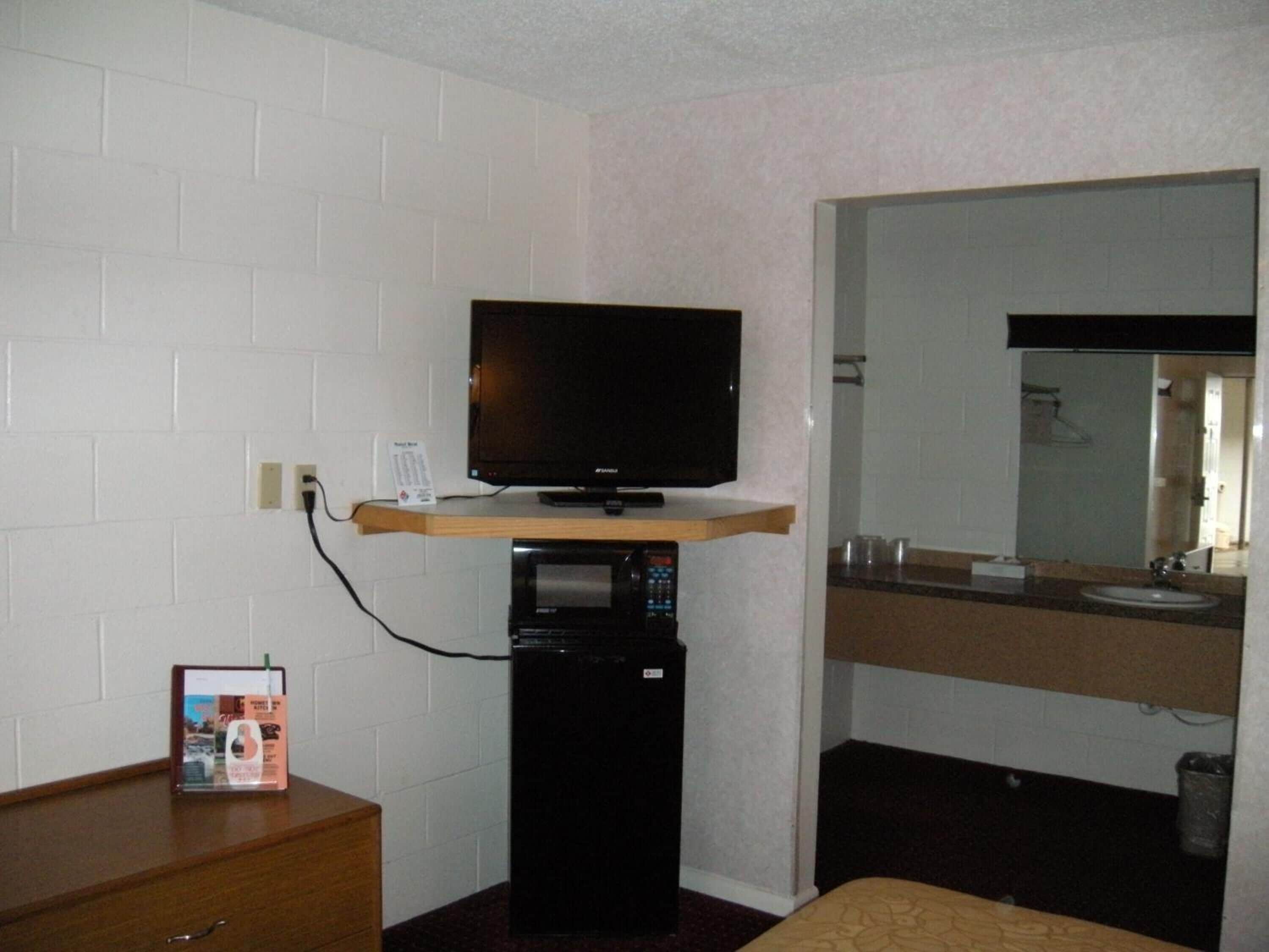 A hotel room with a television on a wooden shelf, a microwave, a small refrigerator, a wooden dresser, a bathroom with a sink and mirror.