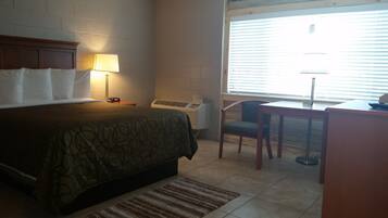 A hotel room with a bed, bedside lamp, air conditioning unit, desk, chair, and a window with blinds.
