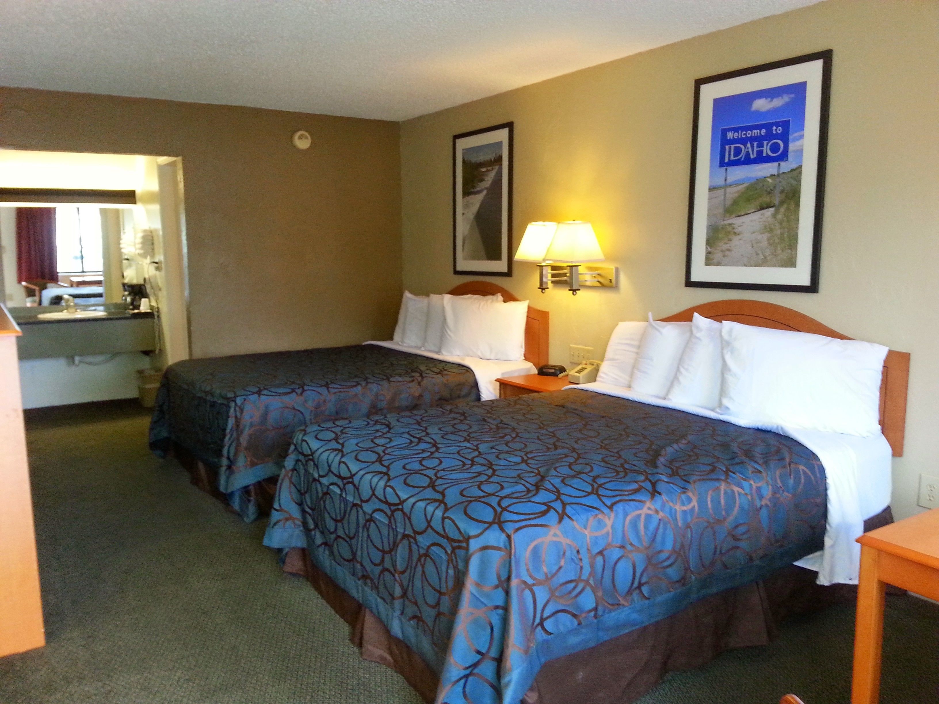 A hotel room with two beds, a nightstand, a lamp, and a framed welcome sign.