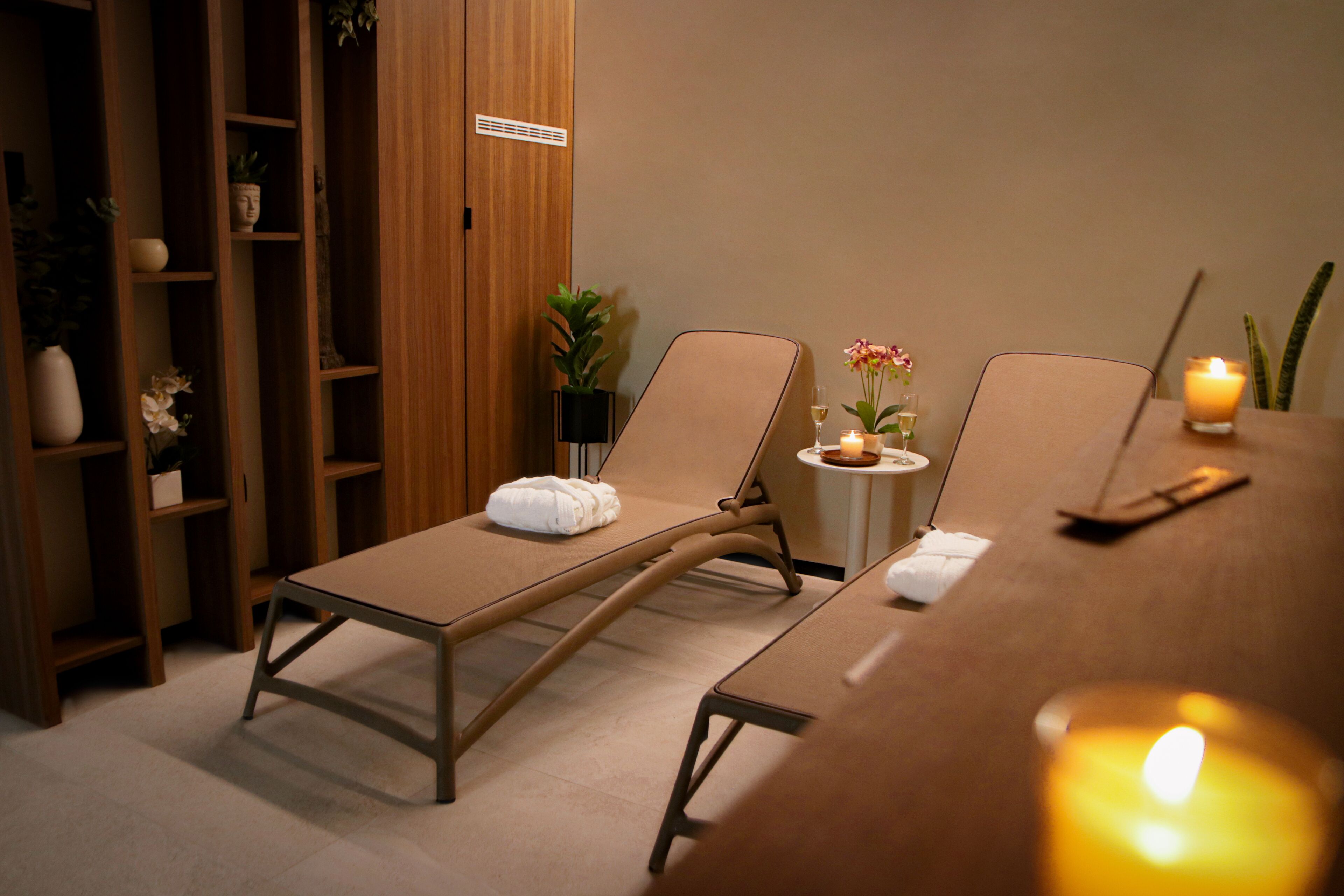 A spa room with two lounge chairs, a small table with candles, and a wooden shelf with plants and decorative items.
