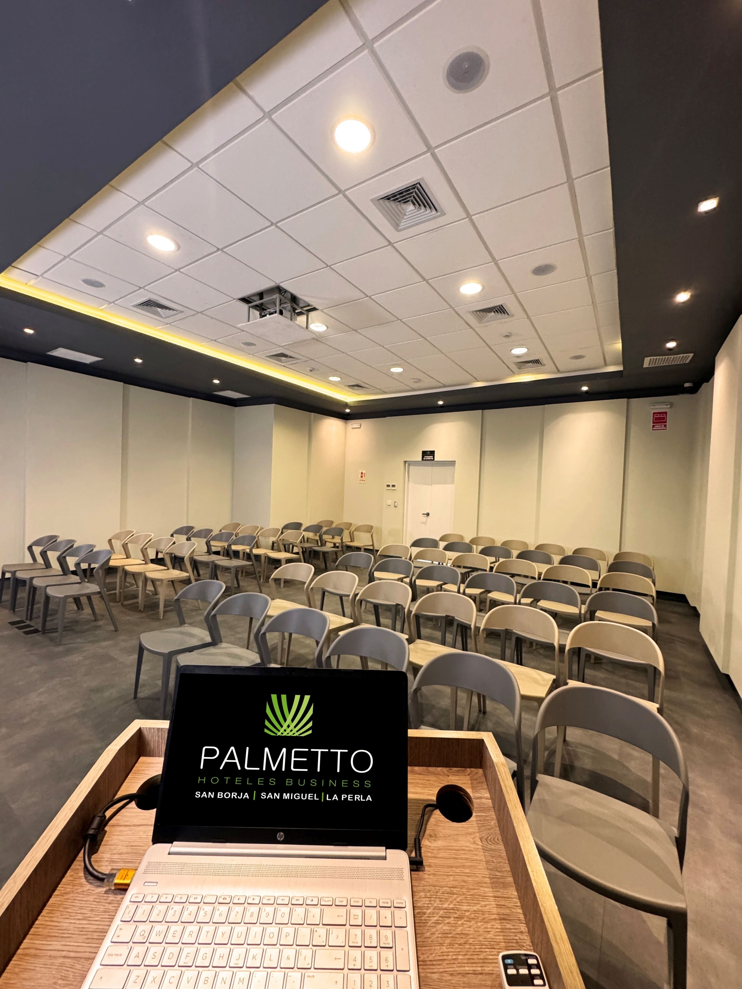 Un salón de conferencias con filas de sillas, una laptop sobre una mesa de madera e iluminación empotrada en el techo.