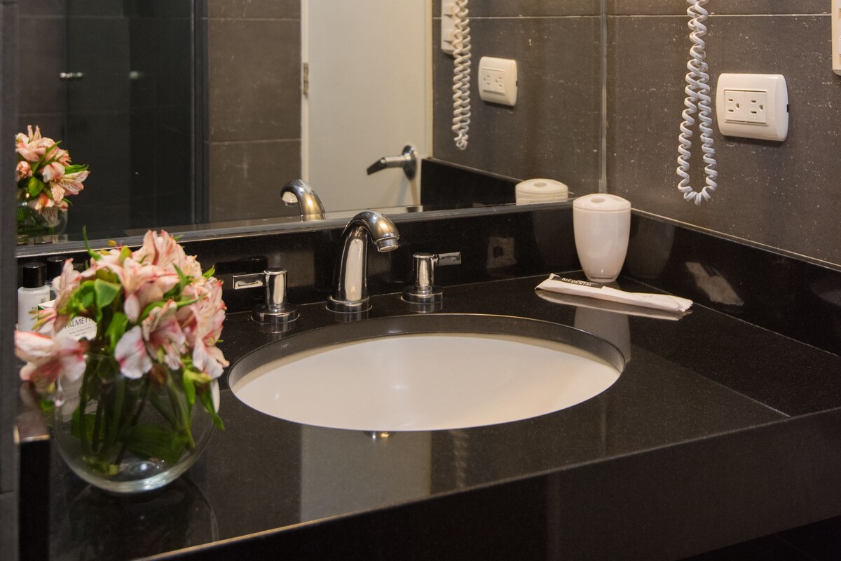 Un baño moderno con encimera negra, lavabo blanco, espejo y secador de pelo montado en la pared.