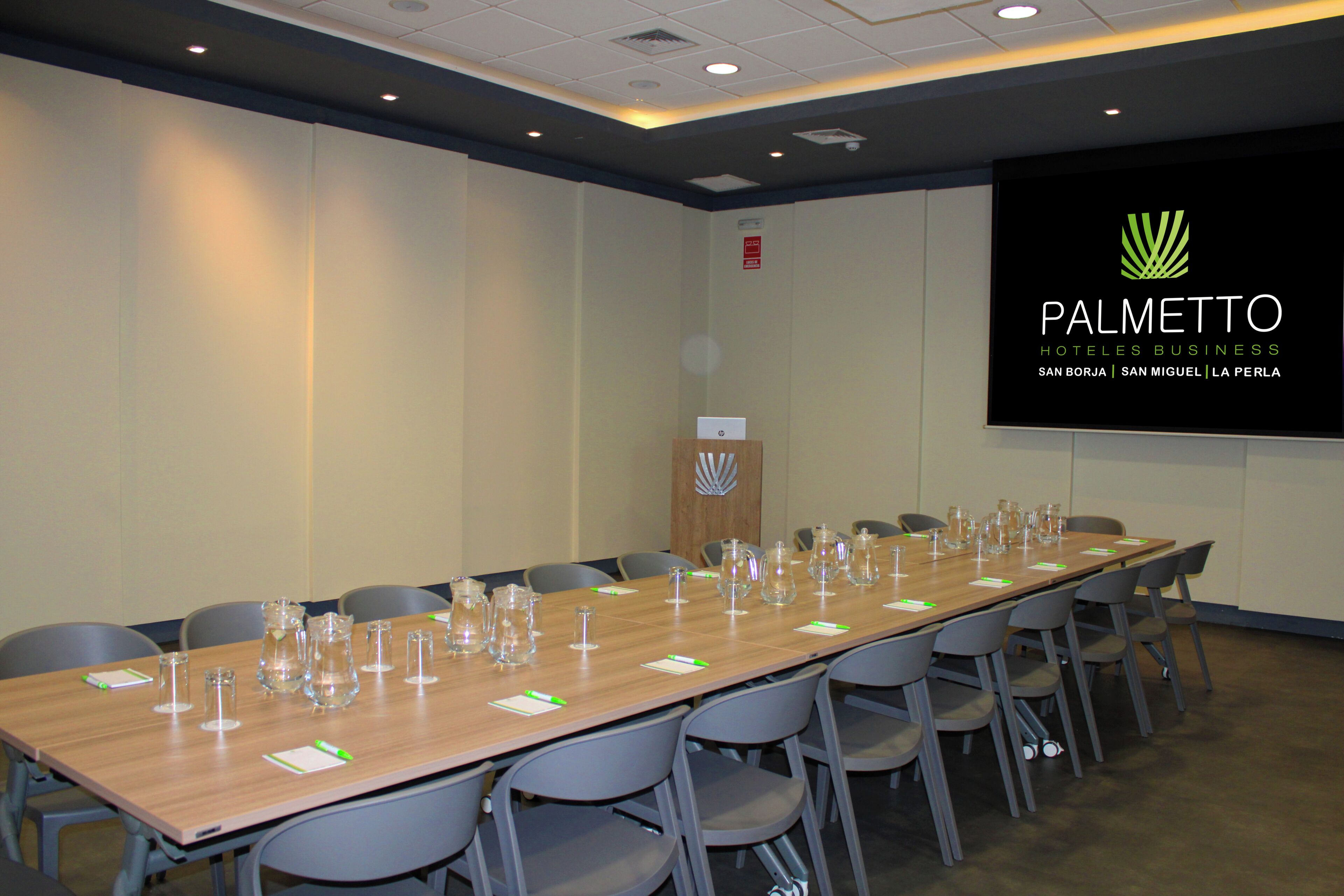 Un salón de conferencias con una mesa larga, sillas y una pantalla mostrando el nombre del hotel.