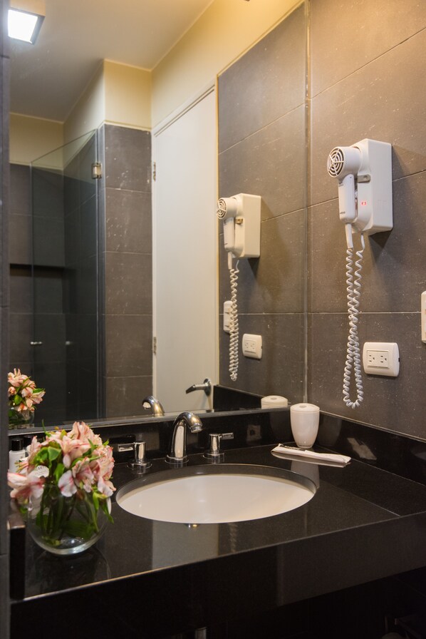 A modern bathroom with a large mirror, a black countertop, a white sink, a hairdryer, and a vase of flowers. - Palmetto Hotel Business San Miguel (Lima)