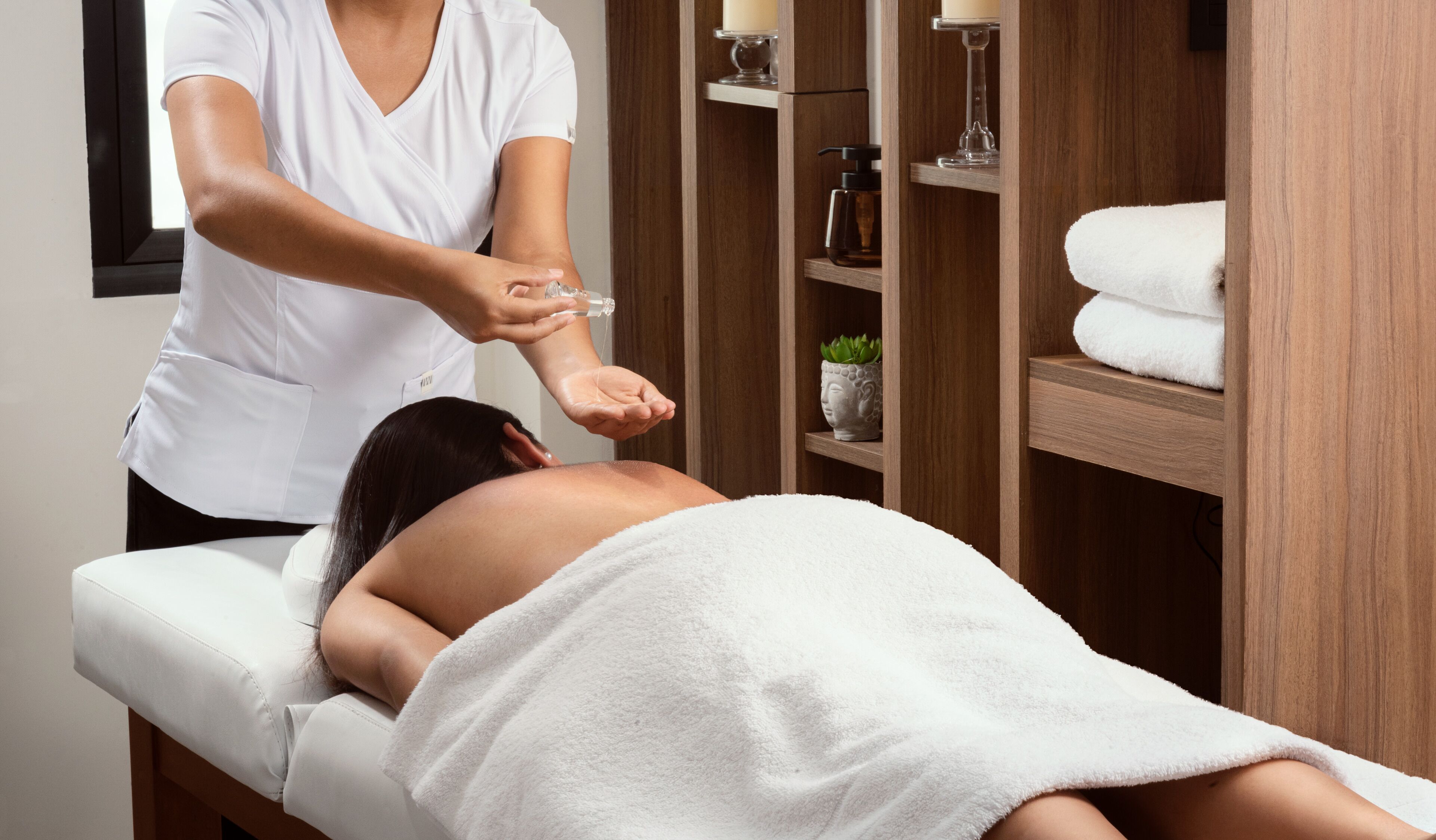 A person receiving a massage in a room with wooden shelves, a white towel, and a small plant.