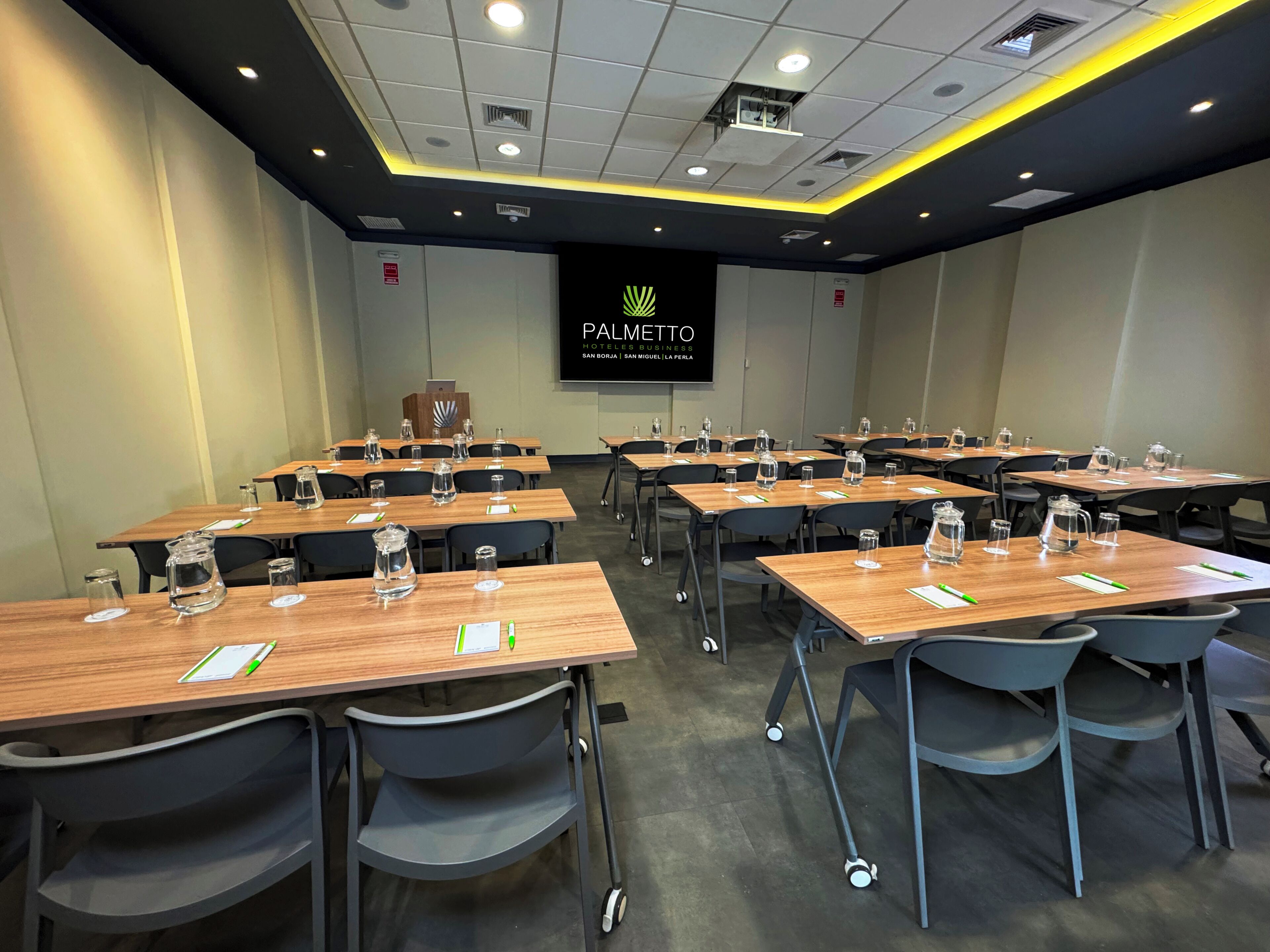 Un salón de conferencias con varias mesas, sillas y una pantalla de proyector que muestra 'PALMETTO HOTEL BUSINESS SAN BORJA | SAN MIGUEL LA PERLA'.