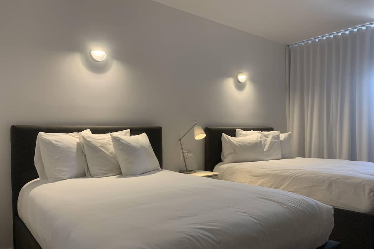 Habitación de hotel con dos camas, cada una con ropa de cama y almohadas blancas, una mesita de noche con lámpara y dos luces fijadas en la pared.