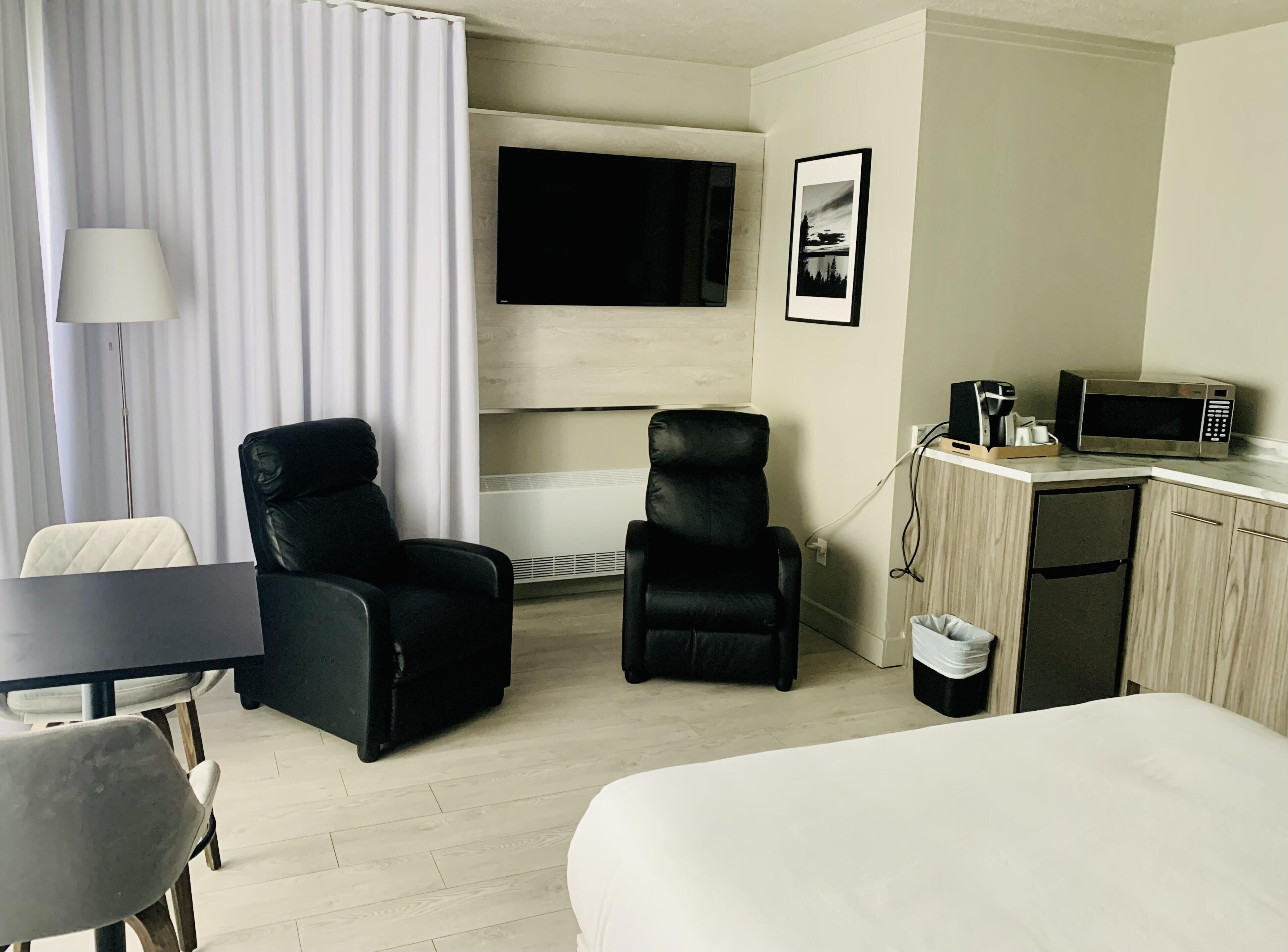 Une chambre d’hôtel comprenant un lit, deux fauteuils en cuir noir, un bureau avec un micro-ondes et un téléviseur fixé au mur.