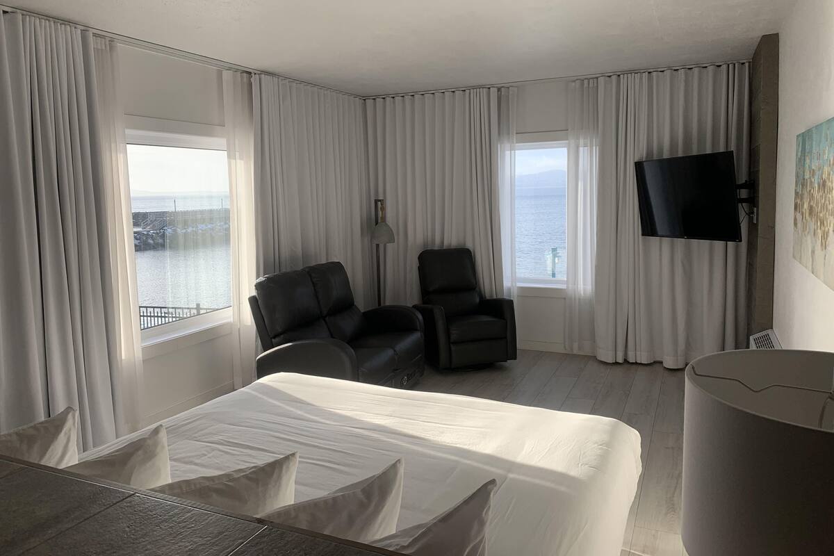 Habitación de hotel con cama, dos sillas, televisor y ventana grande con vistas al mar.