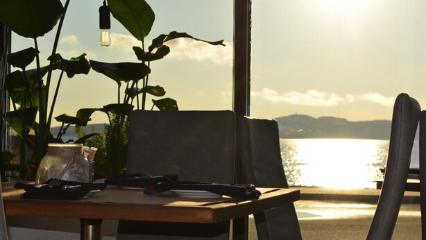 Una mesa con una planta, un tarro y un plato con cubiertos, con vista a un cuerpo de agua y montañas al fondo.