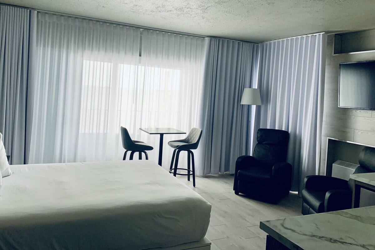 Habitación de hotel con una cama grande, dos sillas, una mesita pequeña, un escritorio, una lámpara y un televisor en la pared.