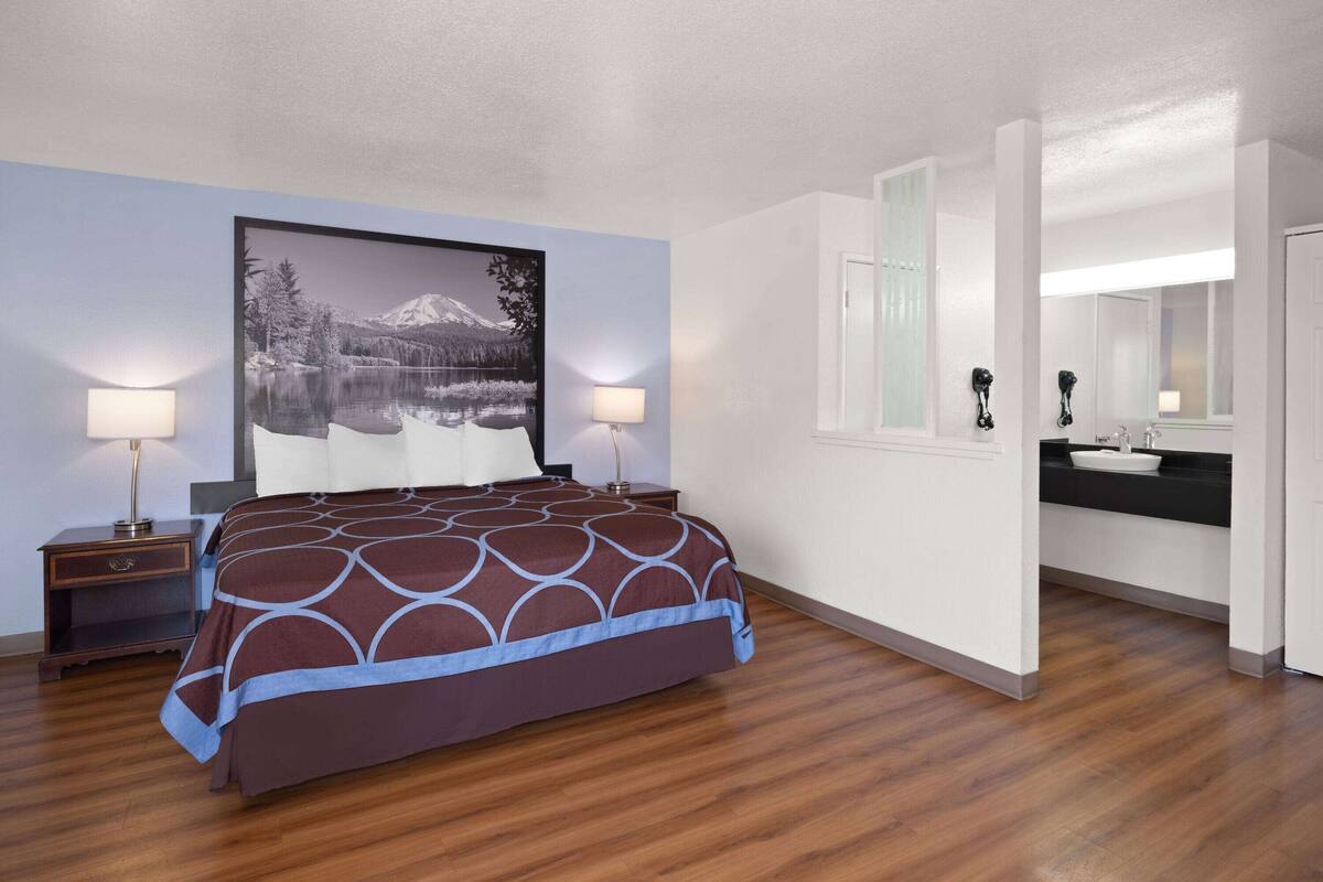 Habitación de hotel con cama, dos lámparas de noche, un cuadro grande en la pared, baño con lavamanos y espejo, y piso de madera.