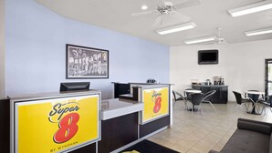 A hotel lobby with reception desks, a ceiling fan, a coffee station, and a wall-mounted TV.