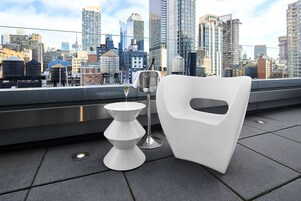 Terraza en la azotea con una silla blanca, una mesita y una copa de vino, con vista a un horizonte urbano con varios edificios.