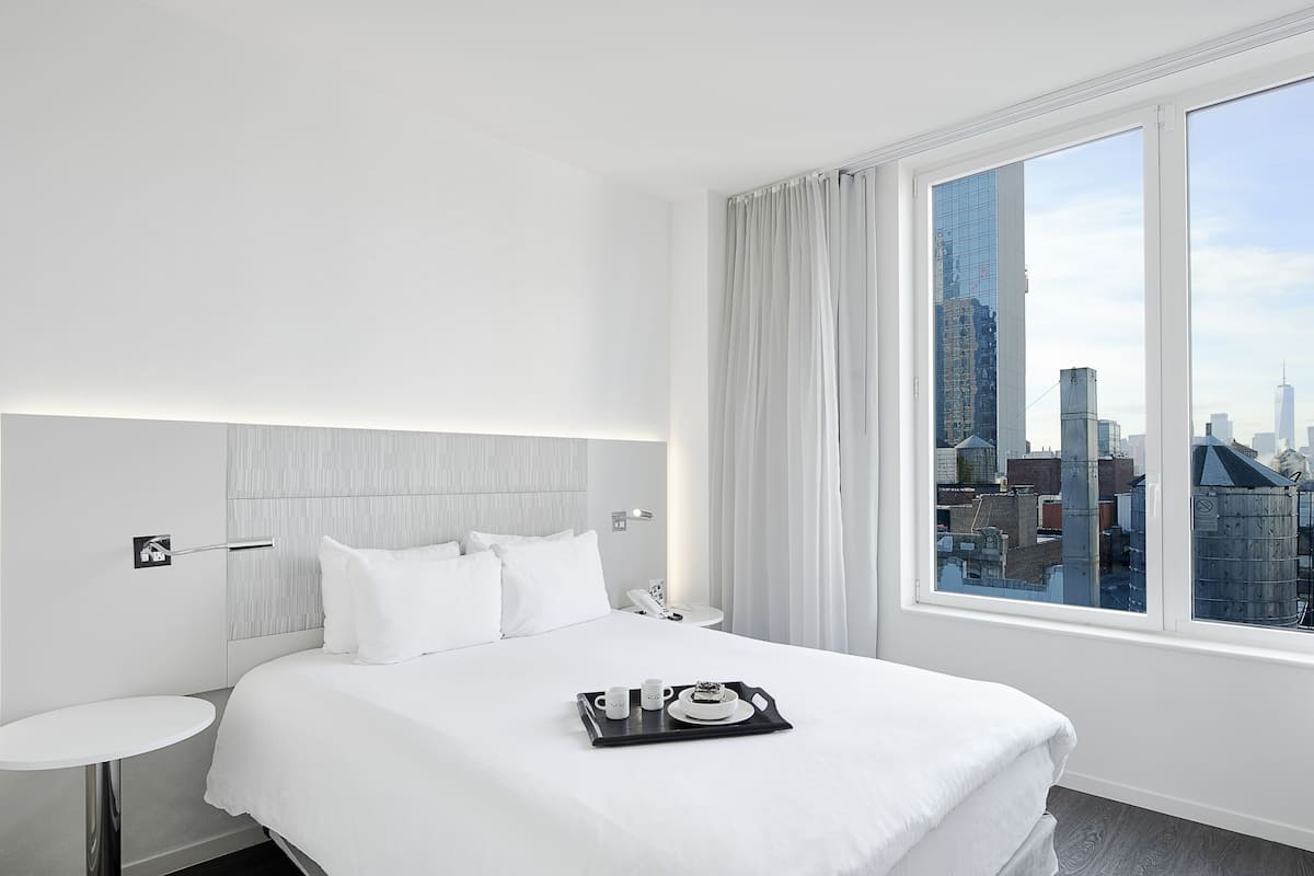 A modern hotel room with a large bed, a bedside table, a small round table, and a window with a city view.